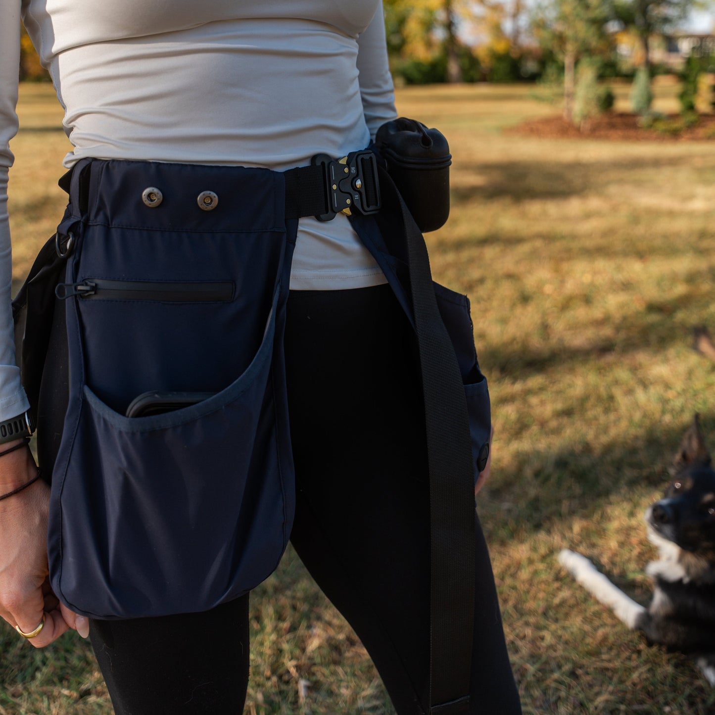 woman wearing a white long-sleeve shirt and dark pants with a blue MALIKO Dog Training belt in an outdoor setting.