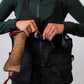 A person wearing a black mesh dog training vest over a dark green long-sleeve top and matching green shorts, adjusting the front zipper and buckle.