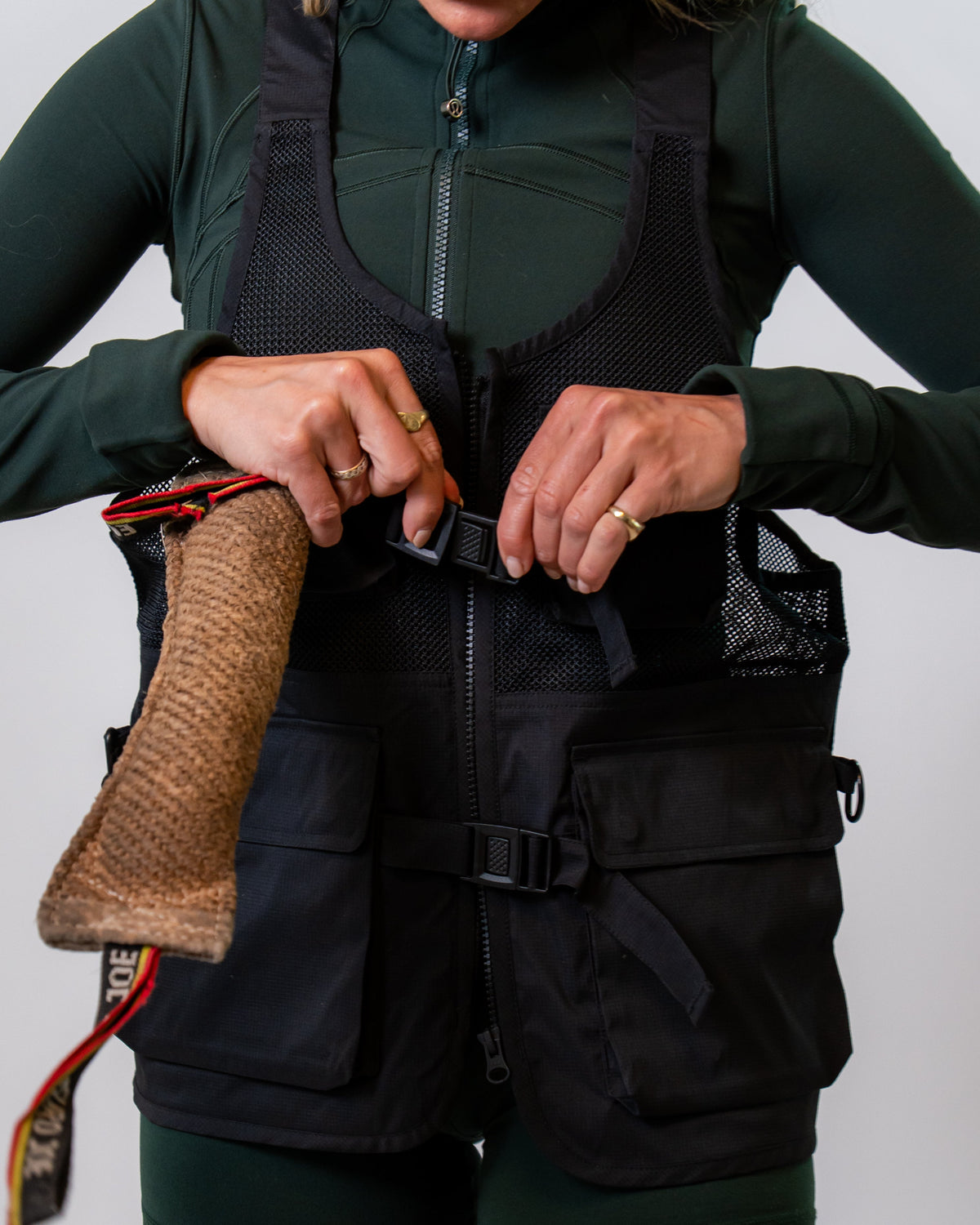 A person wearing a black mesh dog training vest over a dark green long-sleeve top and matching green shorts, adjusting the front zipper and buckle.