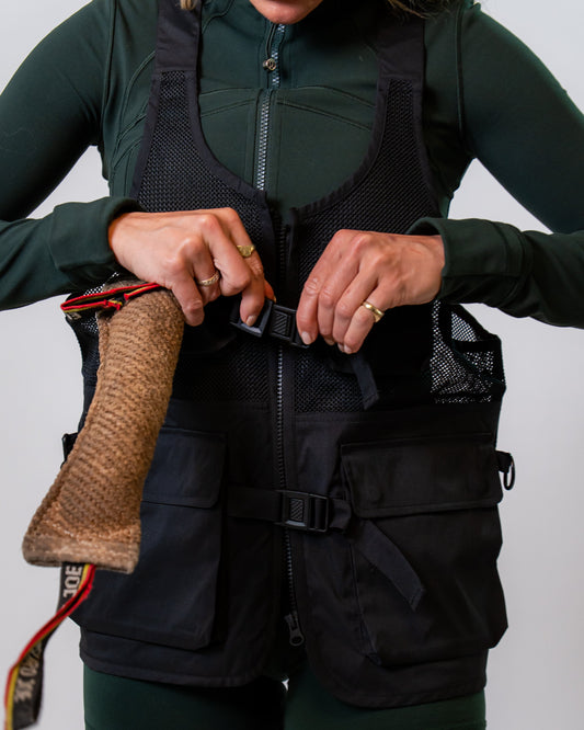 A person wearing a black mesh dog training vest over a dark green long-sleeve top and matching green shorts, adjusting the front zipper and buckle.