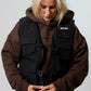 A blonde woman wearing a black MALIKO dog handler vest over a brown hoodie, looking down while adjusting the front buckle. 