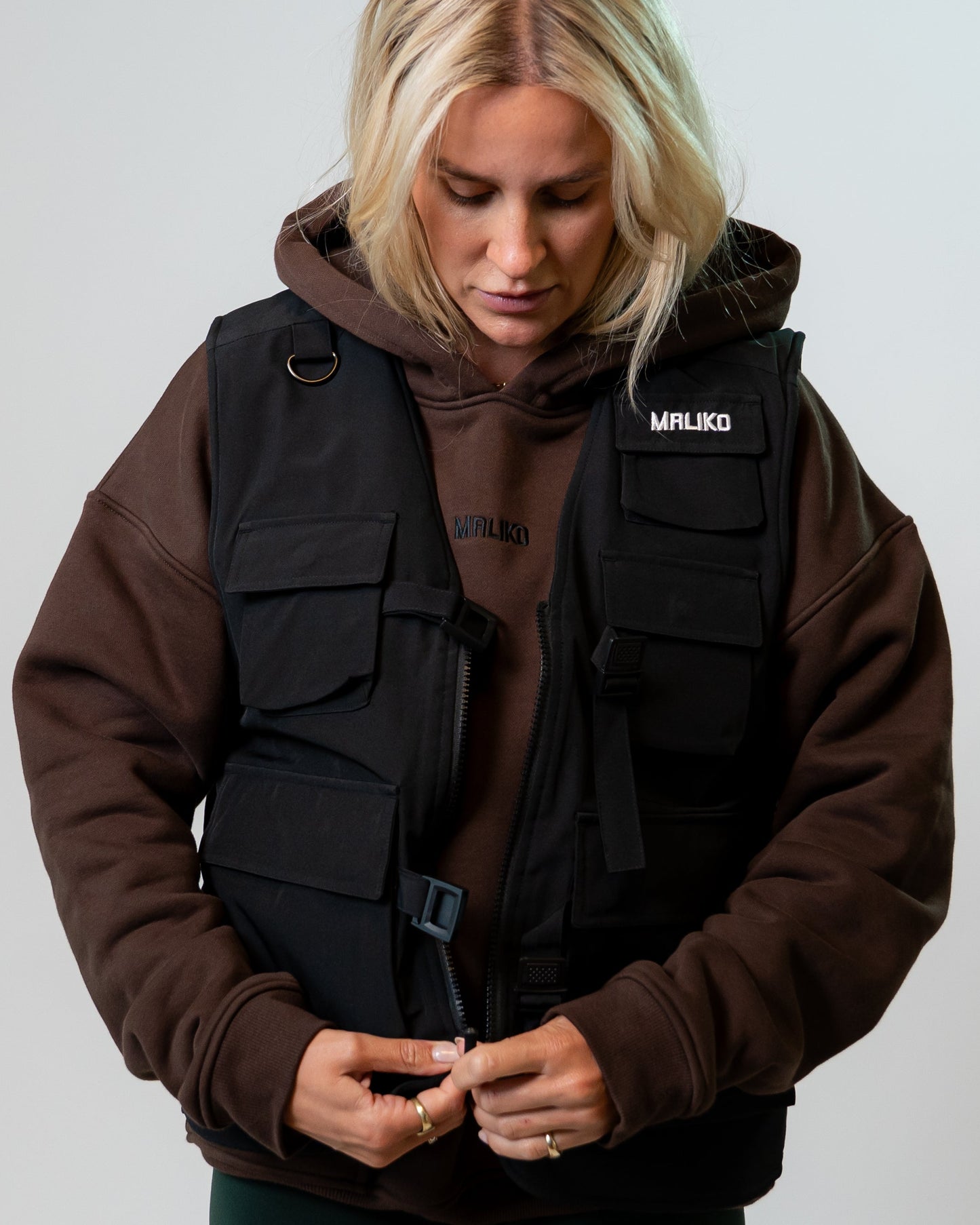 A blonde woman wearing a black MALIKO dog handler vest over a brown hoodie, looking down while adjusting the front buckle. 