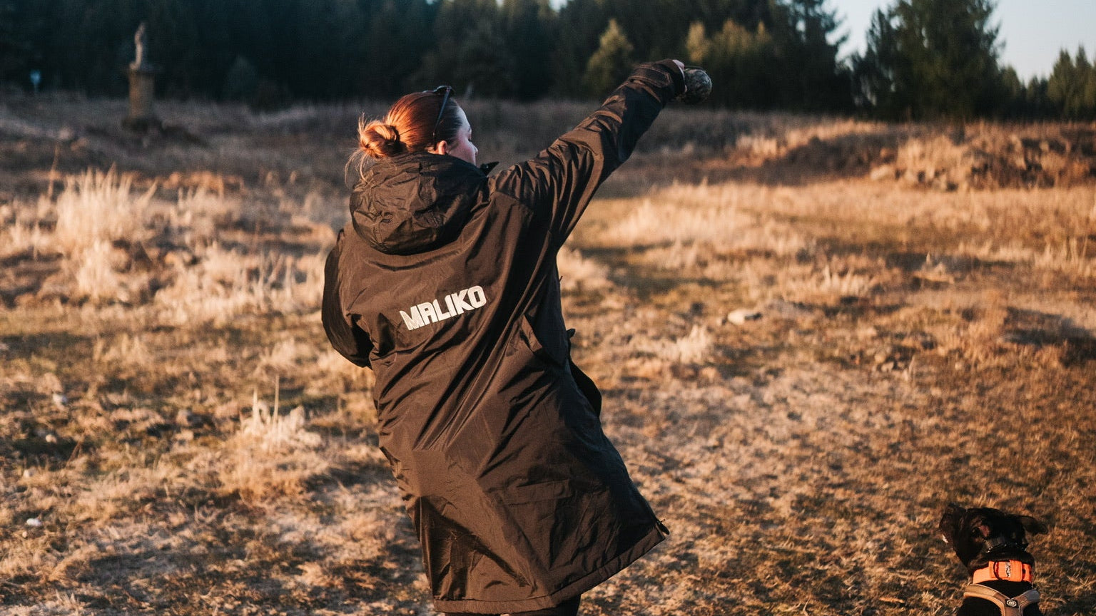 Person in a coat with 'MALIKO' branding playing with a dog in a field.