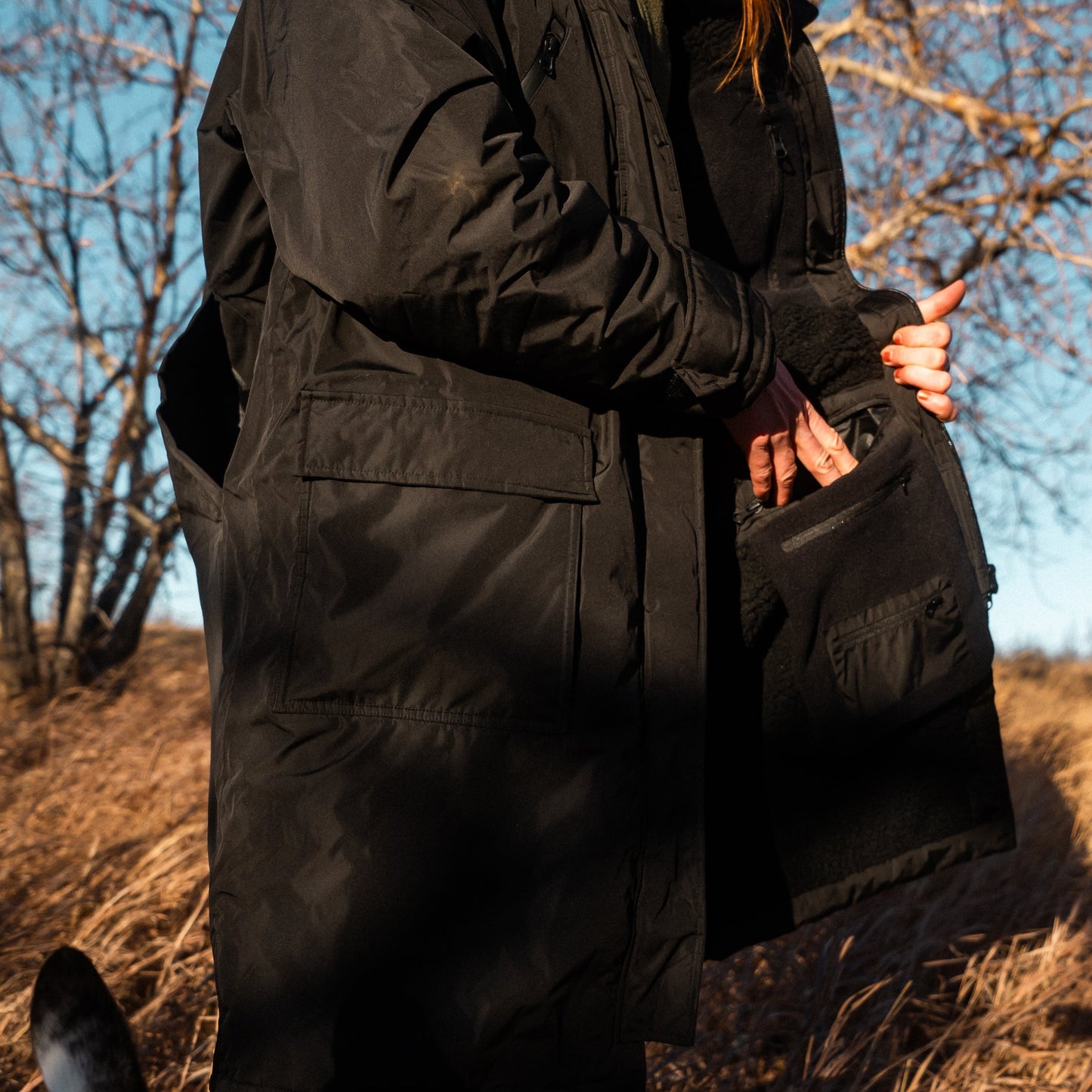 Person in black winter coat and pants standing on a hillside with a dog.