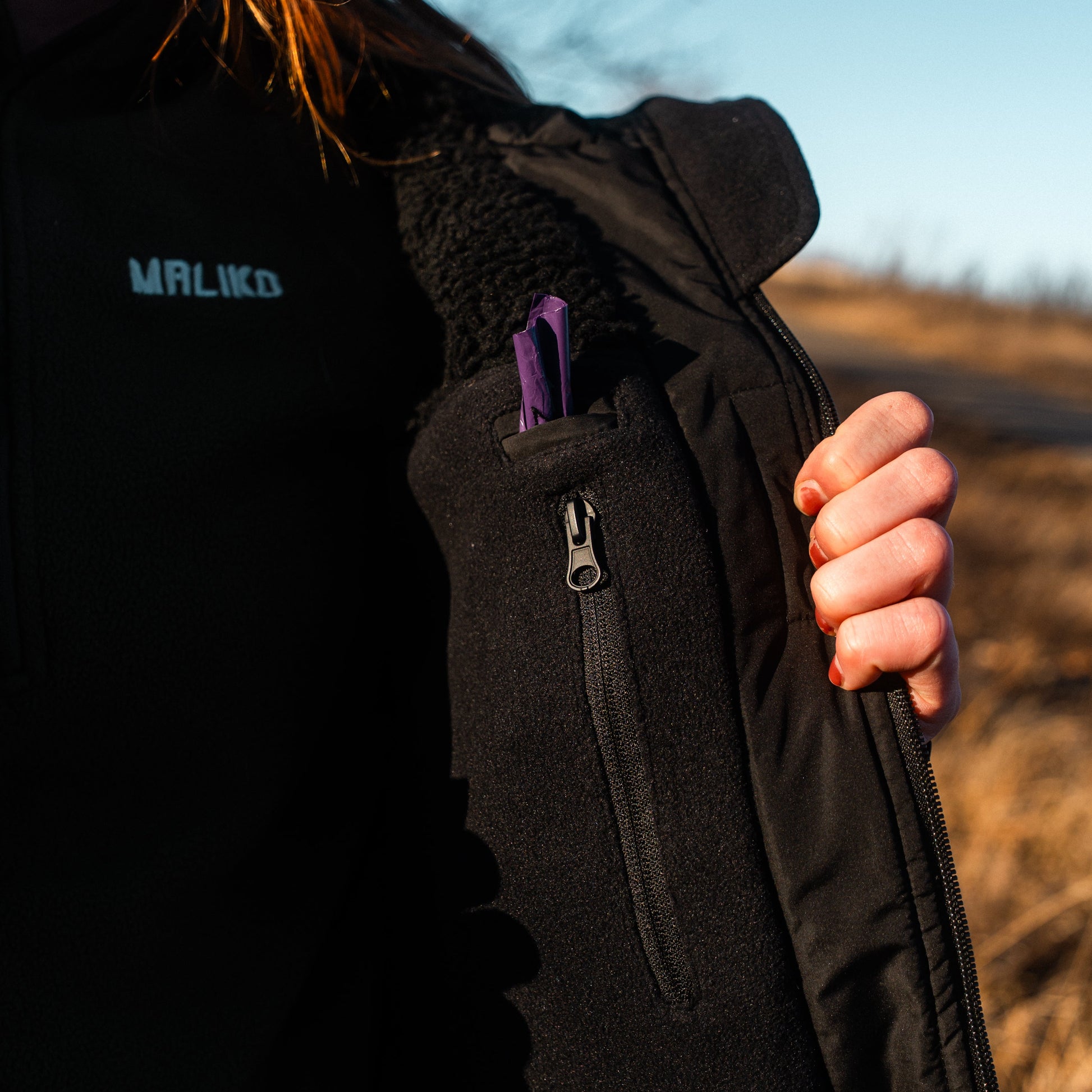 Person wearing a black maliko jacket with a visible brand logo, holding a purple item.