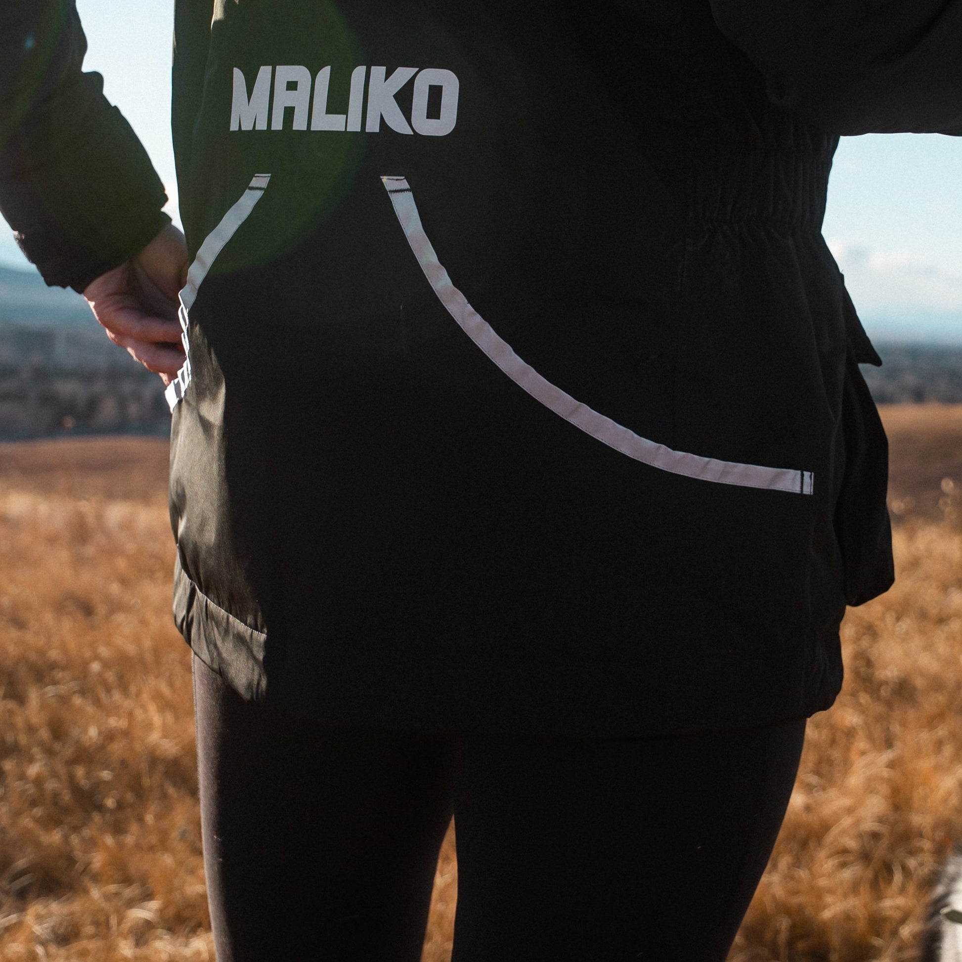 Person wearing a black jacket with 'MALIKO' branding, standing in a field with a dog.