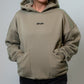 A woman wearing a muted green hoodie with a small black 'MALIKO' logo on the chest, paired with black pants, standing against a plain light gray studio background.