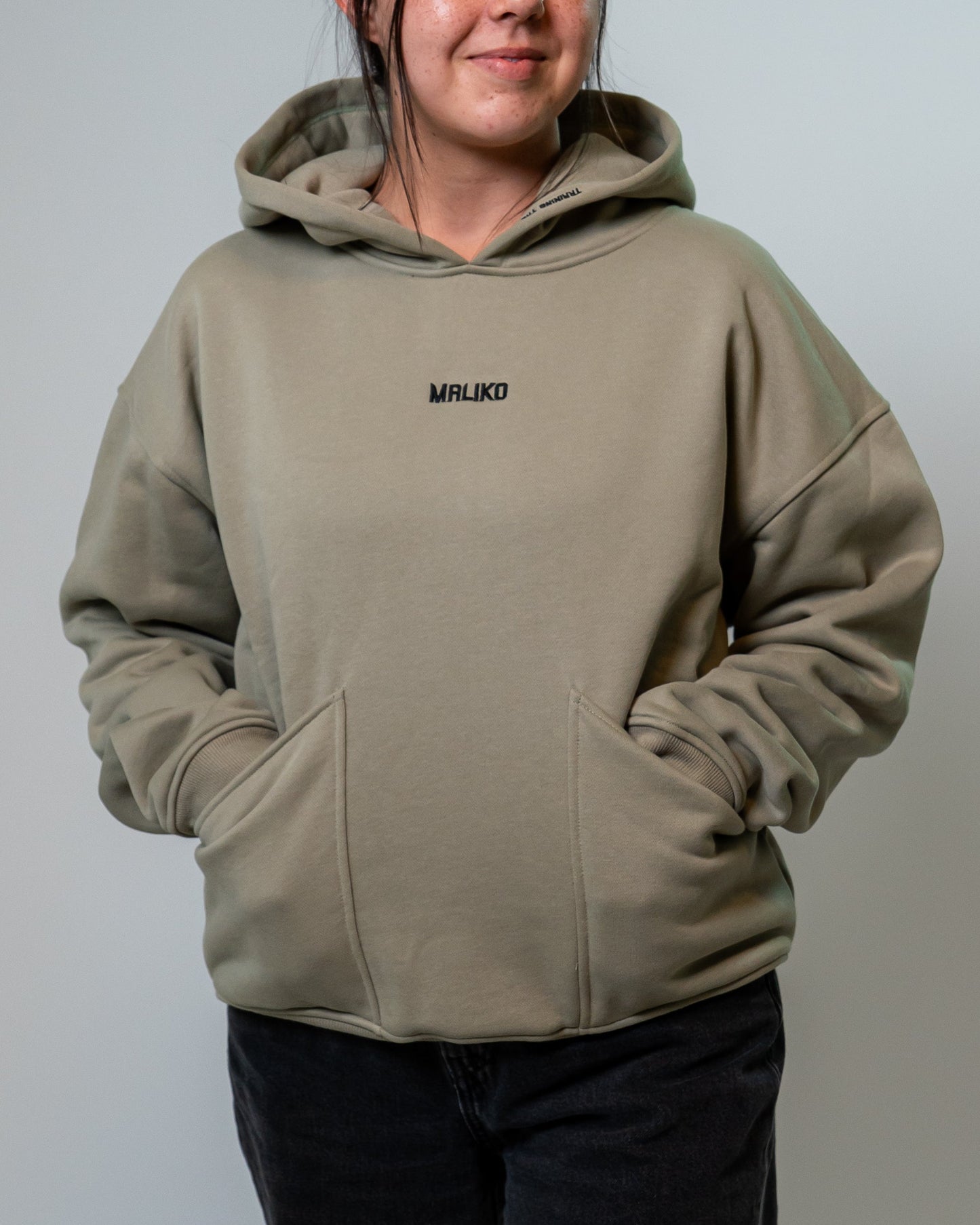 A woman wearing a muted green hoodie with a small black 'MALIKO' logo on the chest, paired with black pants, standing against a plain light gray studio background.