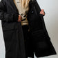 A woman wearing a long black hooded parka over a beige hoodie, holding open the coat to show the interior pockets and lining against a plain light gray studio background.