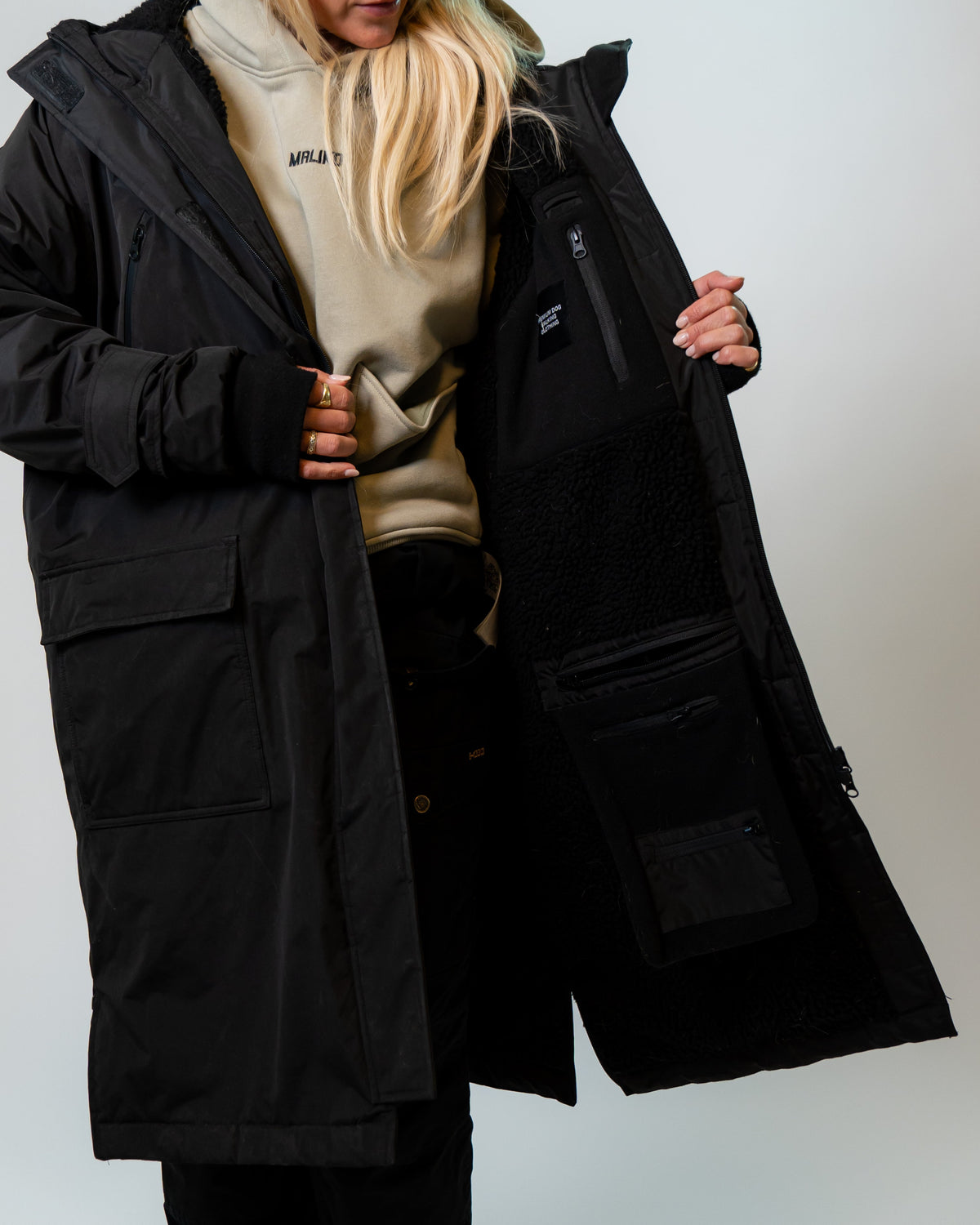 A woman wearing a long black hooded parka over a beige hoodie, holding open the coat to show the interior pockets and lining against a plain light gray studio background.