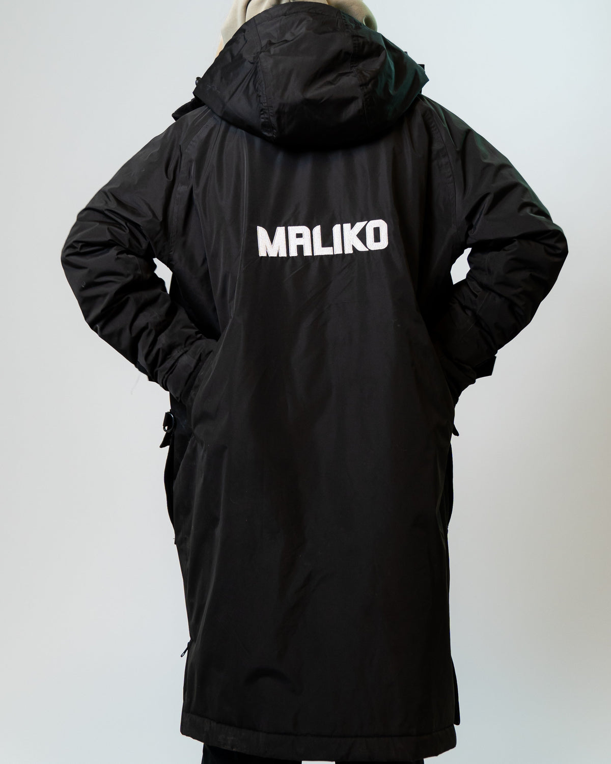 A person viewed from behind wearing a long black hooded jacket with the brand name 'MALIKO' printed in white across the back, against a plain light gray studio background.