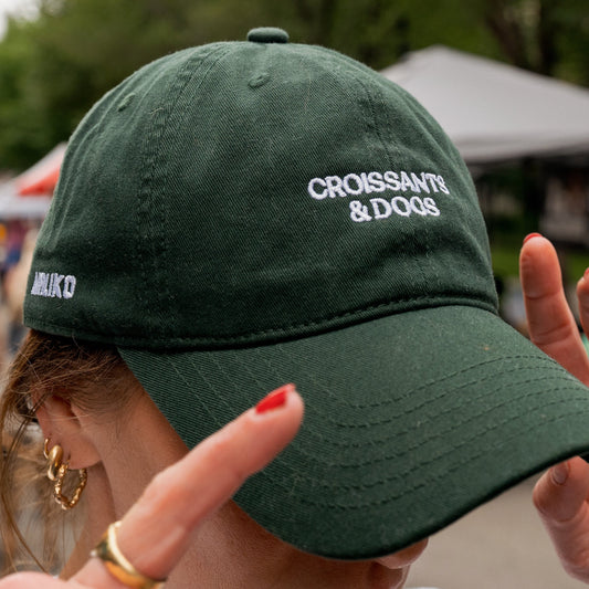 Person wearing a green cap with 'Croissants & Dogs' text, blurred outdoor background