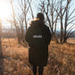 Person wearing a black coat with 'MALIKO' branding walking through a field with bare trees.