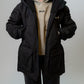 A woman modeling a black hooded puffer jacket over a beige hoodie, standing against a plain light gray studio background.