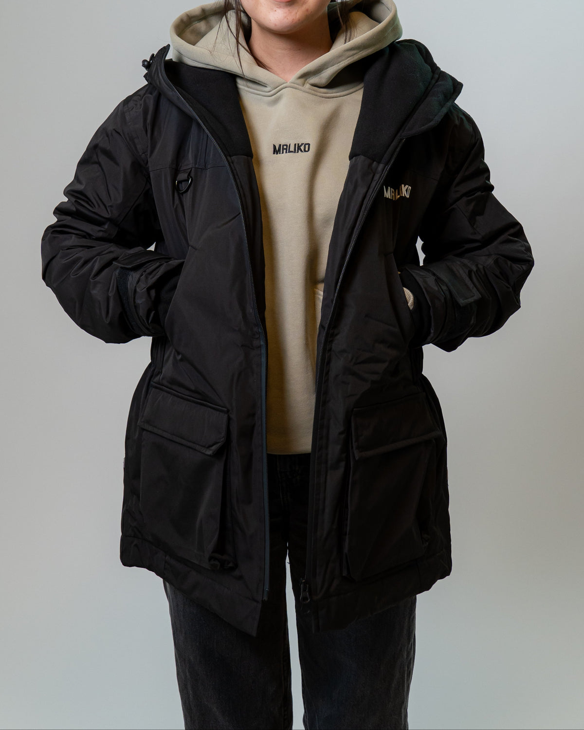 A woman modeling a black hooded puffer jacket over a beige hoodie, standing against a plain light gray studio background.